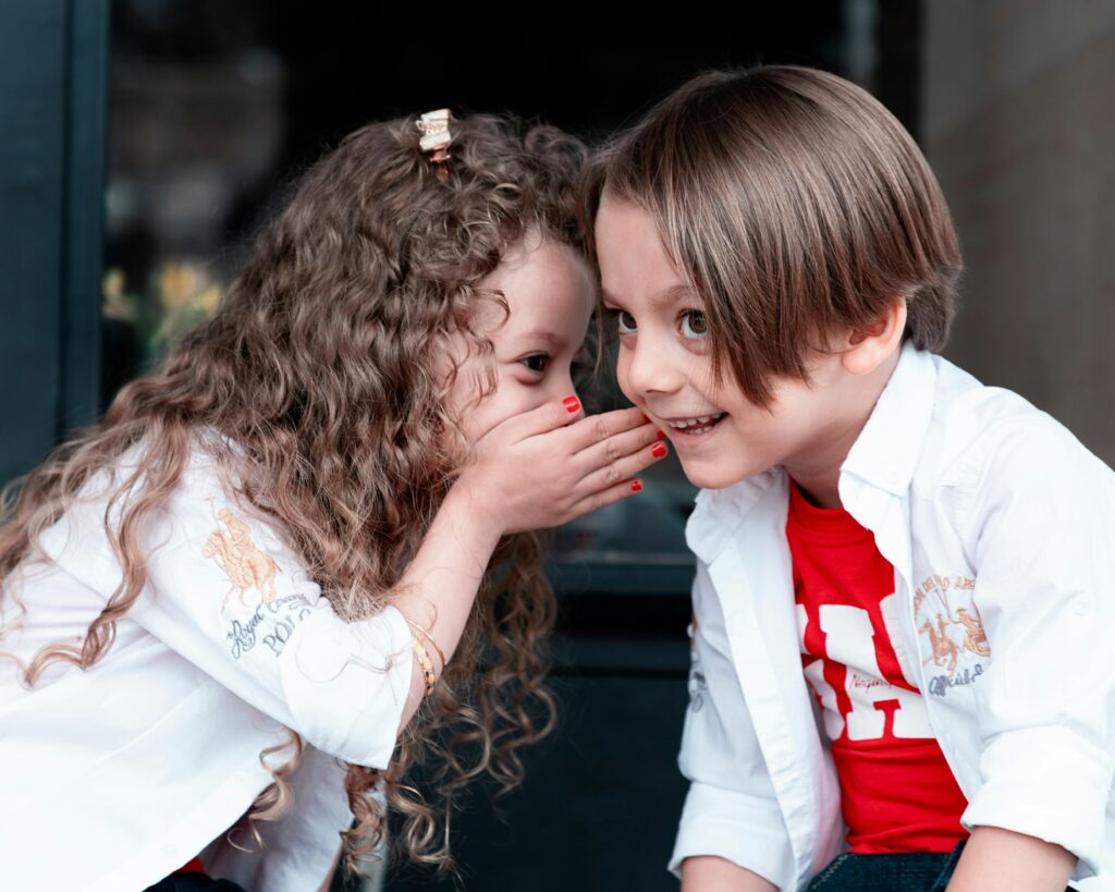 A little girl is whispering to a smiling young boy.